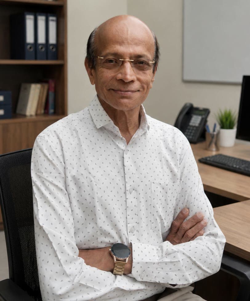 Dr. Mangalore Vittal Kamath, seated at a desk with his arms folded, smiling