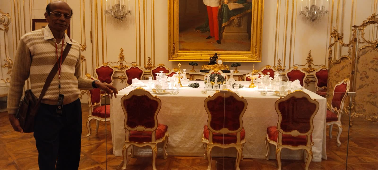 In a gilded palace dining room, beside a long table set with red velvet chairs.