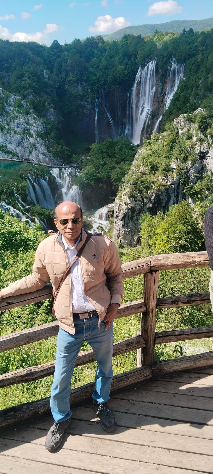 At a wooden lookout rail, with a tall waterfall pouring down green cliffs behind.