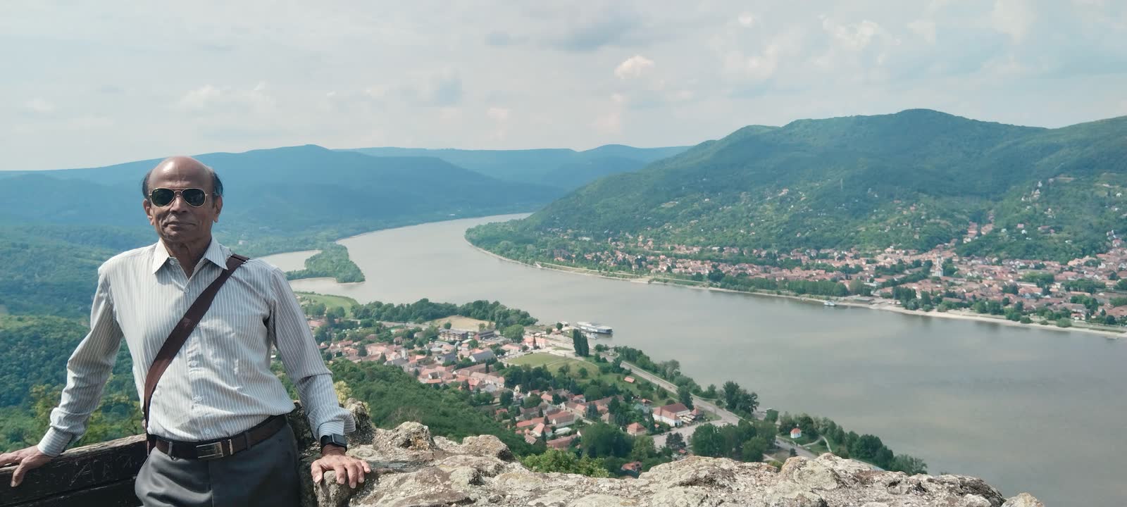 Leaning on a stone wall high above a wide river bend, with a town and wooded hills below.