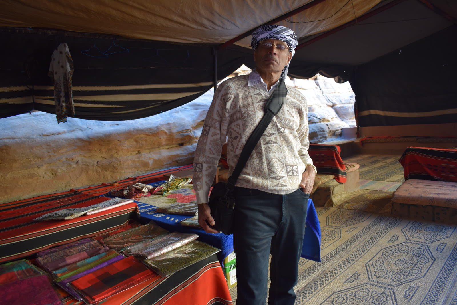 In a patterned sweater and a chequered headscarf, inside a woven tent with rugs and cushions, pale rock beyond the opening.