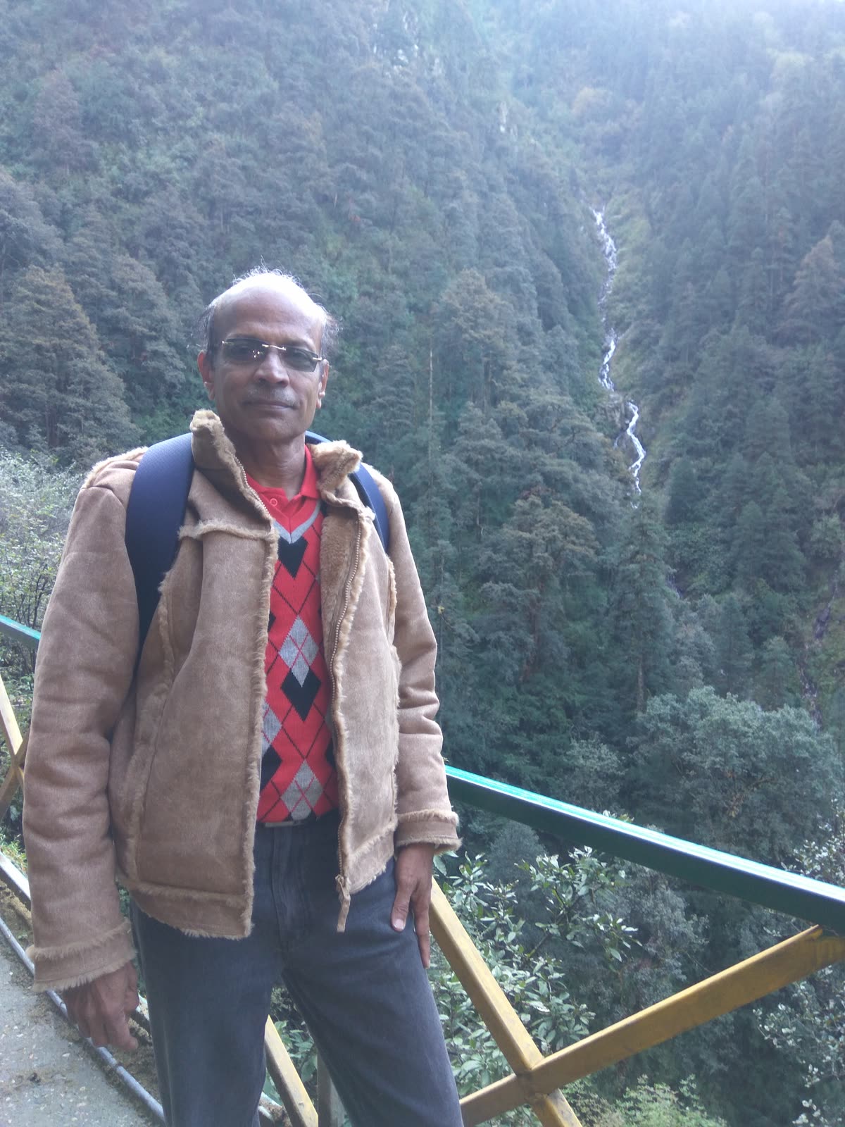 In a sheepskin jacket at a green railing, with forested mountainside and a thin waterfall behind.