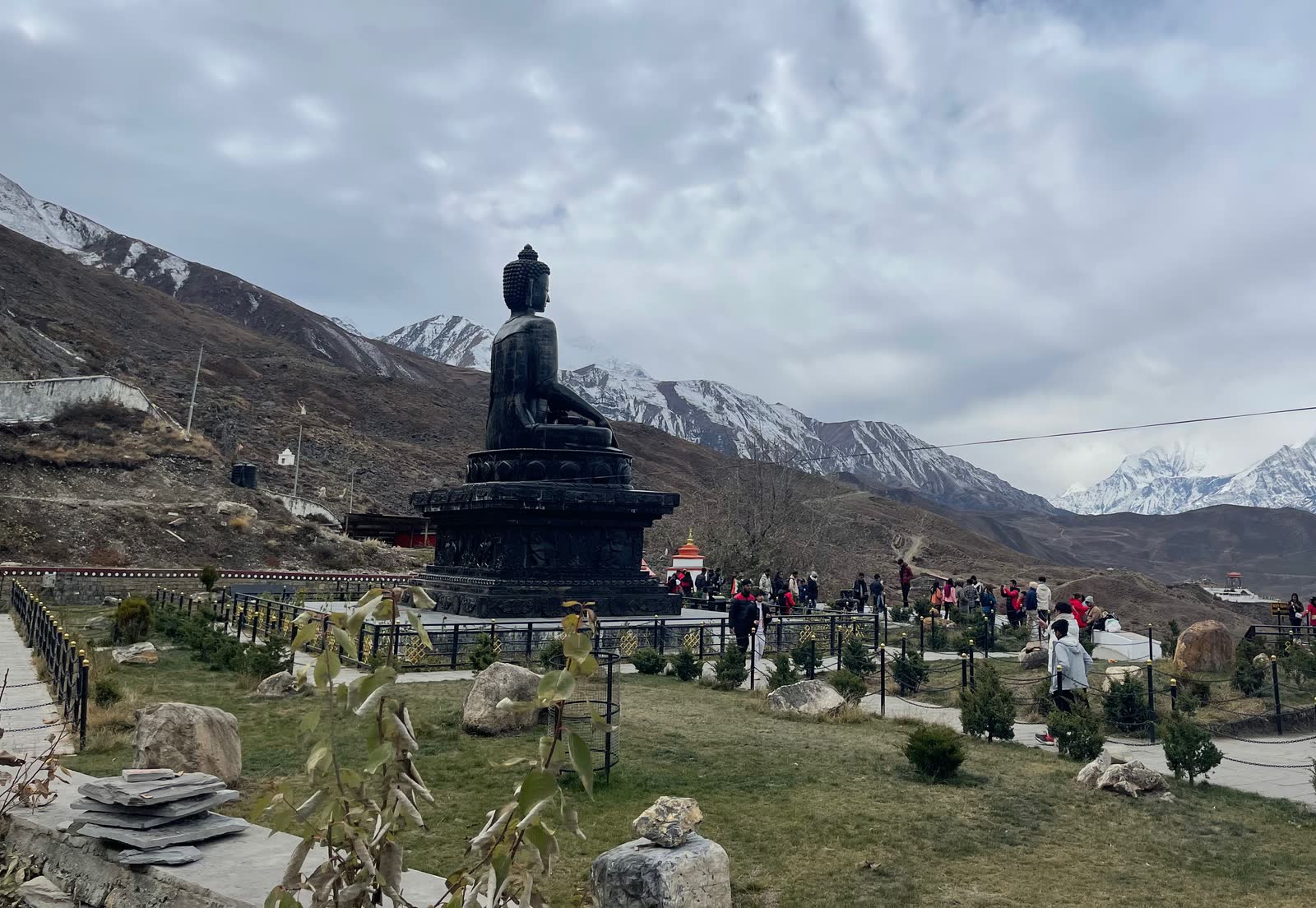 A large dark stone Buddha in the gardens at Muktinath, under cloud, with snow-capped Himalayan peaks beyond.
