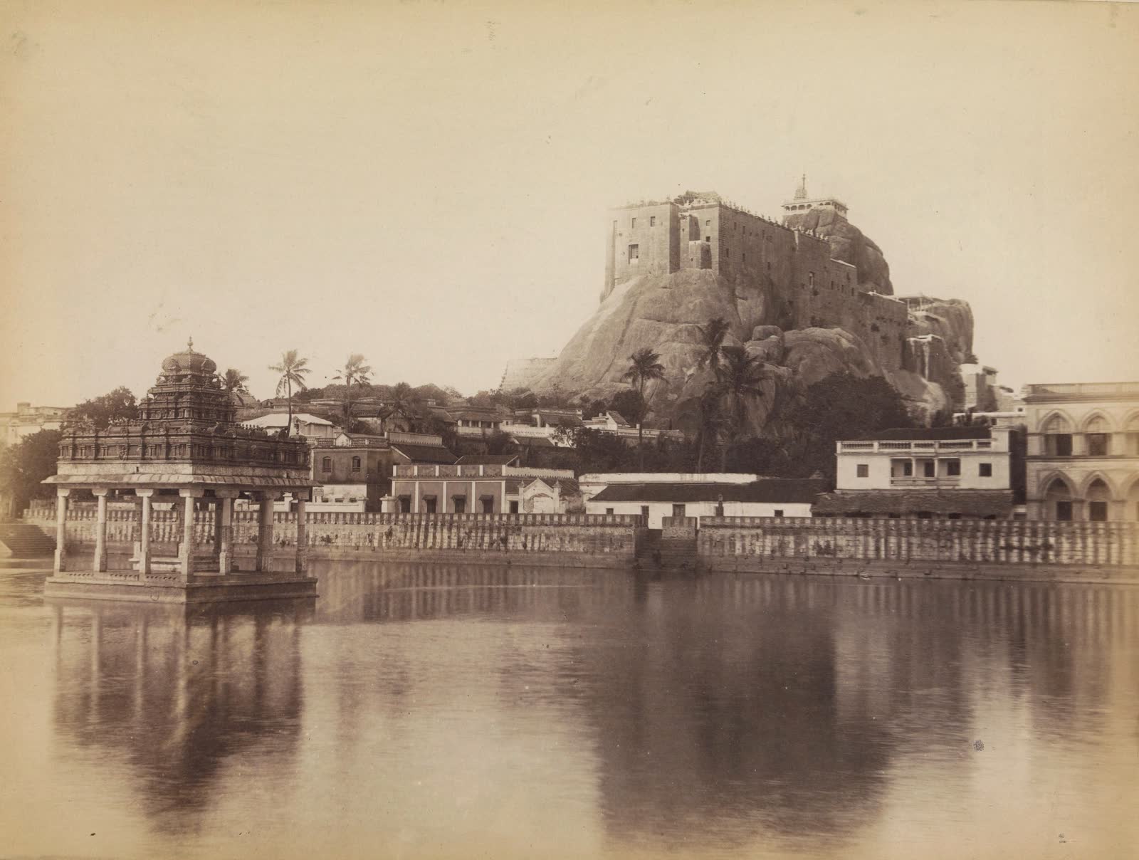 A sepia photograph from about 1870: the fort and temple on the Rockfort rising behind the town, reflected in the temple tank.
