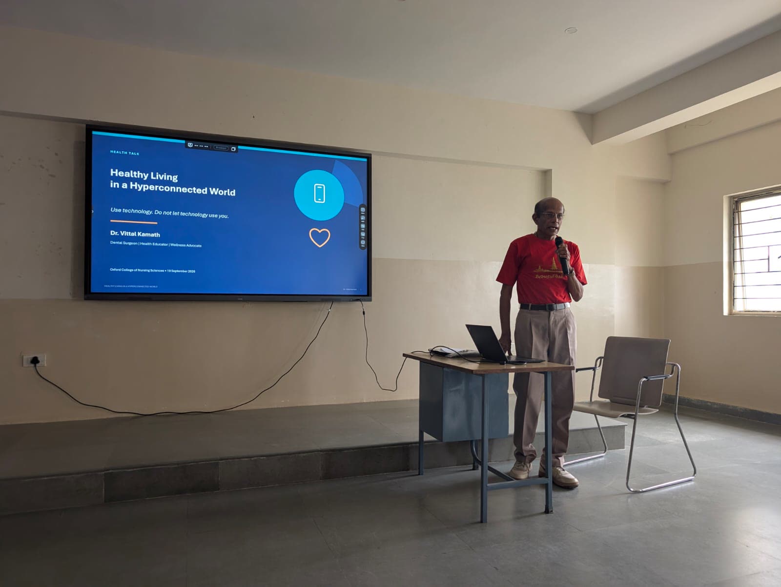 Speaking into a microphone beside a desk and laptop, under a wall screen showing the talk's title slide.