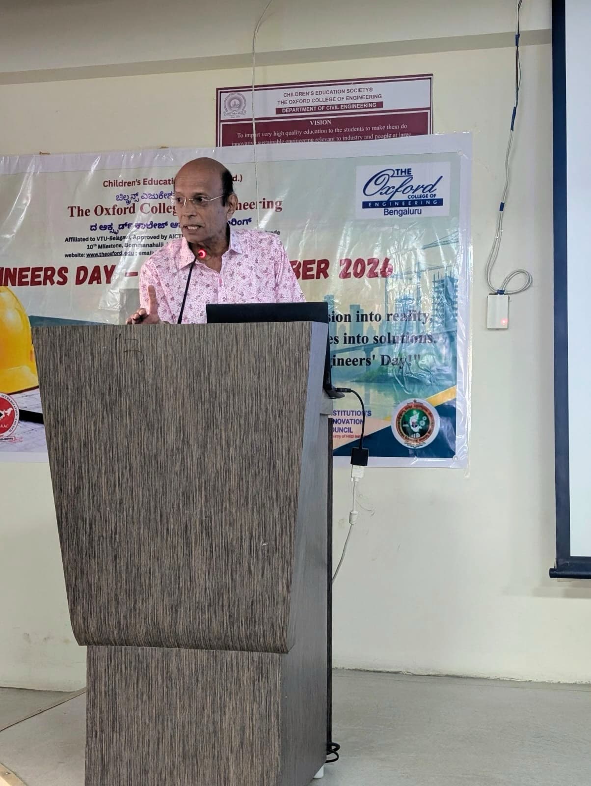 At a wooden lectern with a laptop, one finger raised mid-sentence, in front of a college banner.